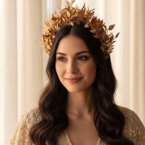 May include: A gold-colored leaf crown, worn on dark wavy hair. The crown is made of multiple leaves and is worn on the head. The person is wearing a gold-colored, embroidered top. The background is a light beige color.