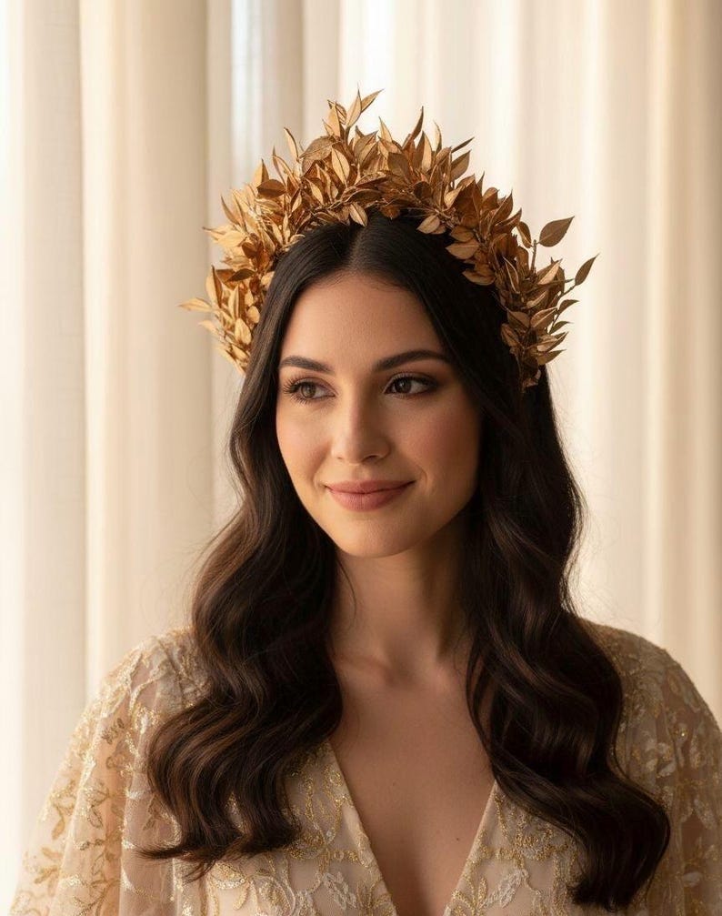 May include: A gold-colored leaf crown, worn on dark wavy hair. The crown is made of multiple leaves and is worn on the head. The person is wearing a gold-colored, embroidered top. The background is a light beige color.