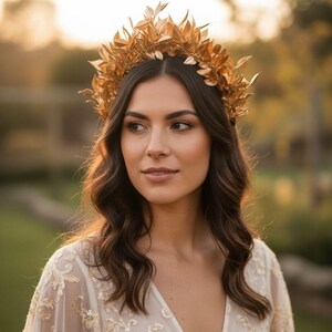 May include: A woman wearing a gold-colored leaf crown. The crown is made of multiple leaves and sits atop dark brown wavy hair. She is wearing a cream-colored dress with floral embroidery. The background is blurred with natural light.