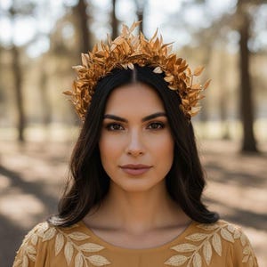 May include: A woman wearing a gold-colored leaf crown and a mustard-colored dress with leaf embroidery. The crown is made of multiple gold leaves. The background is a blurred view of trees.