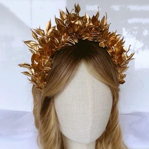 May include: A golden leaf crown, likely for a wedding or special event, sits atop a mannequin head with blonde hair. The crown is made of many small, detailed leaves, creating a full, textured appearance. The leaves are a bright, shiny gold color.