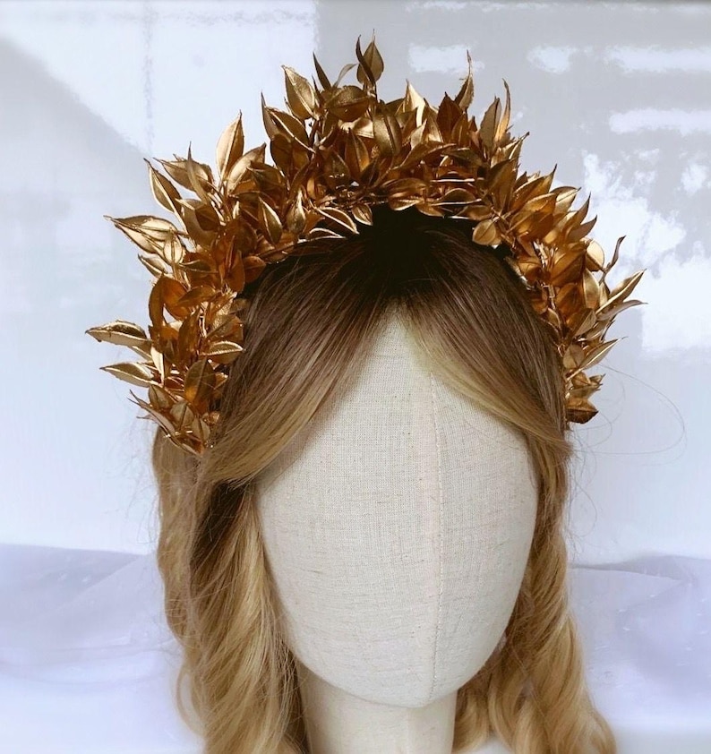 May include: A golden leaf crown, likely for a wedding or special event, sits atop a mannequin head with blonde hair. The crown is made of many small, detailed leaves, creating a full, textured appearance. The leaves are a bright, shiny gold color.