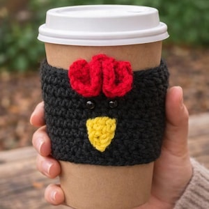 May include: A brown paper coffee cup with a white lid is wrapped in a black crocheted cozy. The cozy features a red rooster comb, black eyes, and a yellow beak. The cup is held by a person.