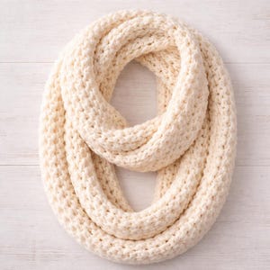 May include: Cream-colored, chunky knit infinity scarf. The scarf is made of a thick, soft yarn and is looped in a circular shape. The knit pattern is visible, creating a textured appearance. The scarf is a neutral color, suitable for various outfits.