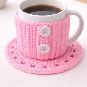 May include: A white ceramic mug filled with dark liquid, encased in a pink crocheted cozy with two white buttons. The mug sits on a matching pink crocheted coaster, creating a coordinated set.
