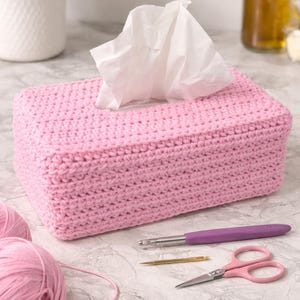 Puede incluir: Una funda rosa de ganchillo para caja de pañuelos con pañuelos blancos. La funda rectangular está sobre una superficie con hilo, un ganchillo, tijeras y una aguja. Tema: decoración del hogar.