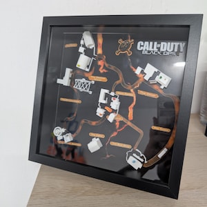 May include: Framed artwork featuring a map from the video game Call of Duty: Black Ops II. The map is a 3D model with white structures and orange pathways against a black background. The frame is black.