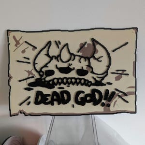 May include: A 3D printed sign with a cartoon skull with horns and the text "DEAD GOD!" on a parchment-like background.