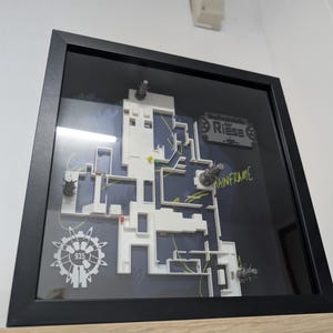 Puede incluir: Un mapa 3D enmarcado con un marco negro. El mapa es blanco con un fondo azul y presenta el texto "Mainframe" y "935". El mapa tiene varios objetos metálicos.