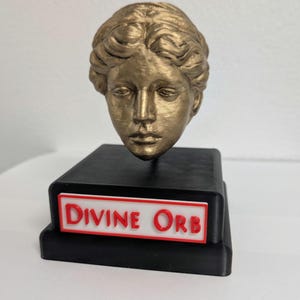Könnte beinhalten: Eine goldfarbene Büste eines klassischen Kopfes auf einem schwarzen rechteckigen Sockel. Der Sockel hat ein weißes und rotes Schild mit der Aufschrift "DIVINE ORB". Die Skulptur ist wahrscheinlich ein dekorativer Gegenstand.