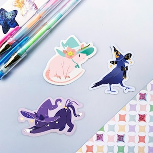 May include: Three witch-themed stickers featuring a pink rat wearing a green and white hat with a flower, a black crow wearing a blue and gold hat with a crescent moon, and a black cat wearing a purple hat with stars.