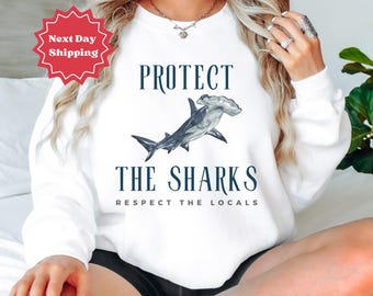 Könnte beinhalten: Weißer Pullover mit dem Text "PROTECT THE SHARKS RESPECT THE LOCALS" und einer Hammerhai-Grafik.