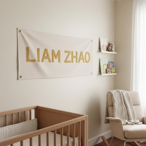 Puede incluir: Una escena de guardería presenta una pancarta con el nombre "LIAM ZHAO" en letras doradas. La pancarta cuelga sobre una cuna y una mecedora. Dos estantes contienen libros y peluches. La habitación tiene una paleta de colores neutros.