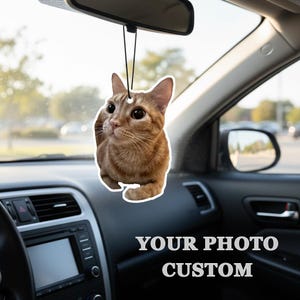Puede incluir: Un ambientador de coche personalizado con un gato atigrado jengibre. El gato cuelga de una cuerda negra, suspendido del espejo retrovisor de un coche. El texto "YOUR PHOTO CUSTOM" es visible en la parte inferior de la imagen.