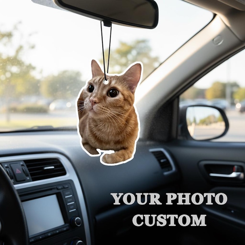 Puede incluir: Un ambientador de coche personalizado con un gato atigrado jengibre. El gato cuelga de una cuerda negra, suspendido del espejo retrovisor de un coche. El texto "YOUR PHOTO CUSTOM" es visible en la parte inferior de la imagen.
