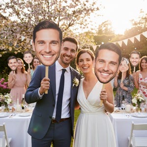 May include: A wedding party poses outdoors, holding large face cutouts on sticks. The bride and groom are in the foreground, smiling. Other guests hold similar cutouts. The scene is set at a table with white linens, decorated with flowers.