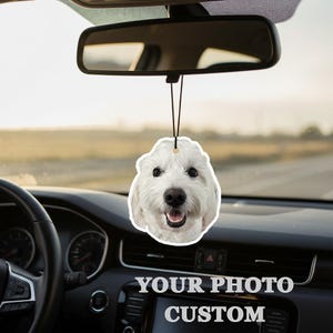 Puede incluir: Un ambientador para coche con forma de cara de perro blanco, colgado del espejo retrovisor de un coche. El ambientador tiene una cuerda negra y una cara de perro sonriente. El salpicadero del coche muestra el texto "YOUR PHOTO CUSTOM".