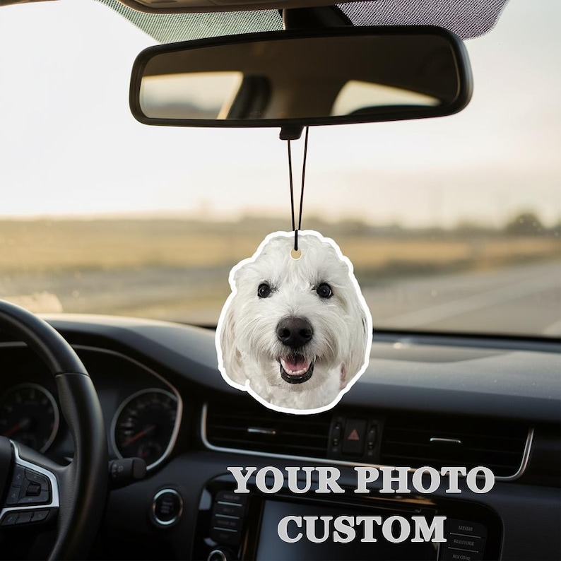 Puede incluir: Un ambientador para coche con forma de cara de perro blanco, colgado del espejo retrovisor de un coche. El ambientador tiene una cuerda negra y una cara de perro sonriente. El salpicadero del coche muestra el texto "YOUR PHOTO CUSTOM".