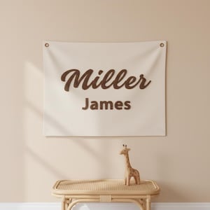 Puede incluir: Un tapiz de pared color crema con el nombre "Miller" y el nombre "James" en escritura marrón. El tapiz de pared tiene dos ojales en la parte superior. Una pequeña jirafa de madera se encuentra en un banco tejido debajo.