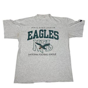 Może przedstawiać: Szary melanżowy t-shirt z krótkim rękawem z ciemnozielonym napisem PHILADELPHIA EAGLES oraz dużym logo NFL poniżej, zawierającym ilustrację orła. Napis NATIONAL FOOTBALL LEAGUE znajduje się w dolnej części nadrukowanej grafiki.