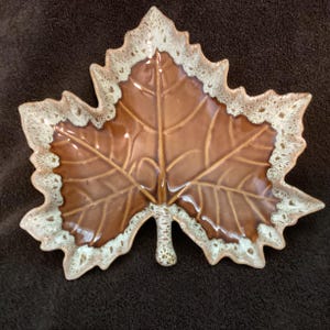 May include: A ceramic dish shaped like a maple leaf with a glossy brown center and a textured, speckled cream-colored border. The surface features embossed vein details that radiate from the center stem toward the jagged, pointed edges of the leaf.