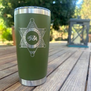 May include: Olive green insulated tumbler with a silver rim and base. The tumbler features a silver emblem with the text "DEPUTY SHERIFF LOS ANGELES COUNTY" and a bear graphic.