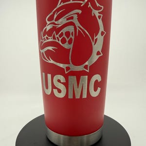 May include: A red insulated tumbler with a silver bulldog graphic and the letters "USMC". The tumbler has a silver rim at the top and bottom and a clear lid. The design is etched into the red surface.