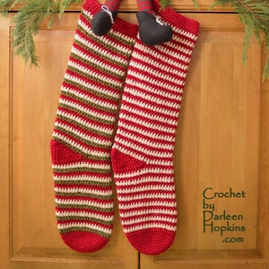 May include: Two red and white striped crocheted Christmas stockings with green accents hanging on a wooden door. A small elf doll is sitting on top of the stockings.