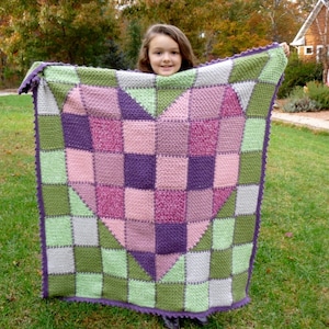 May include: A crocheted heart-shaped blanket in shades of green, pink and purple. The blanket is being held by a young person.