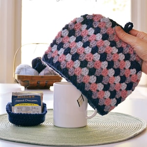 May include: A crocheted tea cozy in shades of pink, blue, and gray. The cozy is being placed over a white mug with a tea bag inside. The mug is sitting on a green placemat. The text "Tea for ME!" is written below the image.