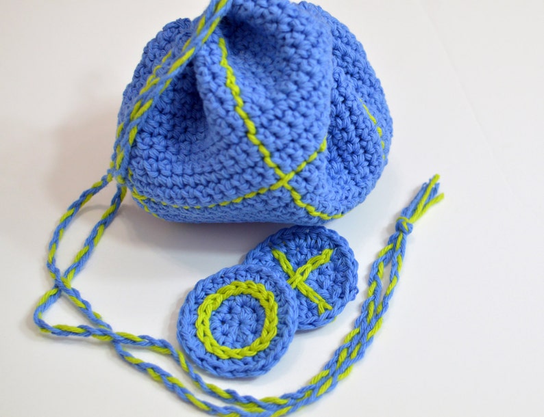 Puede incluir: Una bolsa de cord&oacute;n de crochet azul con ribete verde y dos c&iacute;rculos de crochet a juego. La bolsa tiene un cord&oacute;n largo que tambi&eacute;n est&aacute; tejido en crochet azul y verde.