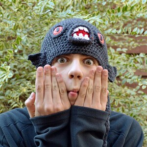 Può includere: Una persona indossa un cappello all'uncinetto nero con un designo a forma di viso di mostro. Il cappello ha occhi rossi, denti bianchi e una lingua bianca e nera. La persona sta facendo una smorfia divertente con le mani che le coprono le guance.