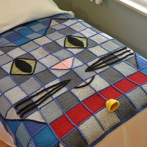 May include: A handmade, colourful cat-themed blanket. The blanket features a large cat face design with blue, grey, red, and yellow squares. The cat's eyes are green and black, and the nose is pink. A small yellow hat rests on the blanket.