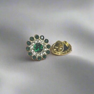 Puede incluir: Un pin de tono dorado con una joya verde rodeada de joyas más pequeñas y transparentes.