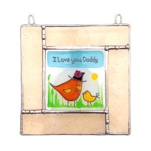 Puede incluir: Una ventana de vitral colgante con un fondo beige claro y un panel central azul y blanco. El panel central presenta un pájaro amarillo con un sombrero rosa y negro con el texto "Te quiero papá".