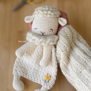 Puede incluir: Una manta de seguridad de cordero de ganchillo, de color crema. El cordero tiene una cabeza esponjosa, orejas pequeñas y una cara sonriente. Una flor amarilla está bordada en la manta.