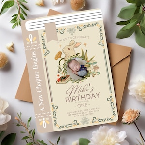 May include: A beige and green illustrated book-shaped invitation with a white rabbit and a baby sleeping in a wreath of flowers. The text reads "A New Chapter Begins", "Join Us To Celebrate", "Mike's Birthday", "As He Turns - One -", "November 21, 2023 - 5:00 PM", "123 Anywhere St, Any City", "RSVP - 123-456-7890".