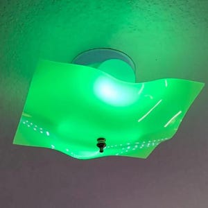 May include: A modern ceiling light fixture with a vibrant green, wavy, square shade. The fixture has a circular base and a small, dark-colored decorative element at the bottom of the shade. The light emits a bright green glow.