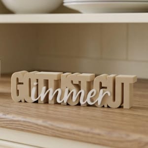 Könnte beinhalten: Ein beigefarbenes Schild in Blockschrift mit dem Text GOTT IST GUT und dem Wort immer in weißer Schreibschrift, das mittig darübergelegt ist. Das Schild besteht aus dickem, festem Material und steht aufrecht auf einer ebenen Fläche.