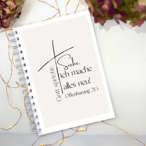 Könnte beinhalten: Ein spiralgebundenes Notizbuch mit cremefarbenem Einband. Der Einband zeigt ein schwarzes Kreuz und den deutschen Text "Gott spricht: Siehe, ich mache alles neu! Offenbarung 21,5". Das Notizbuch ist von Lichterketten und Trockenblumen umgeben.