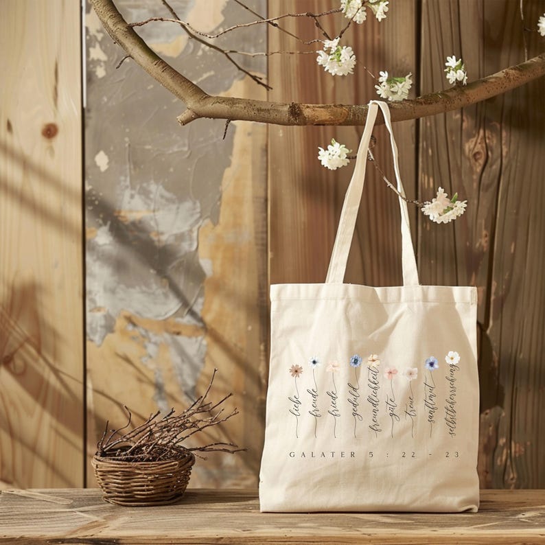May include: A cream-colored tote bag with a floral design and German text. The bag hangs from a tree branch with white flowers. The text reads "Liebe, Freude, Friede, Geduld, Freundlichkeit, G&uuml;te, Treue, Sanftmut, Selbstbeherrschung" and "GALATER 5: 22-23".