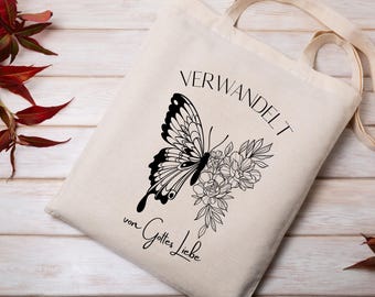 Könnte beinhalten: Eine beigefarbene Stofftasche mit einer schwarzen Strichzeichnung eines Schmetterlings und Blumen. Der Text "VERWANDELT" ist über dem Schmetterling gedruckt, und der Text "von Gottes Liebe" steht in schwarzer Schrift unter dem Blumenmotiv.