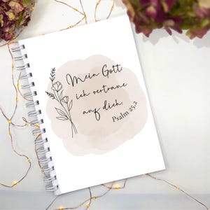 Könnte beinhalten: Ein weißes Notizbuch mit Spiralbindung und dem Text "Mein Gott, ich vertraue auf dich. Psalm 25,2" in schwarzer Schrift. Auf der linken Seite des Textes befindet sich eine florale Illustration. Der Hintergrund ist ein weicher, beiger Aquarell-Wasch.
