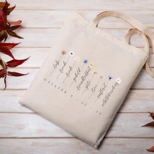 May include: Beige canvas tote bag with a floral design and German text. The bag features a series of watercolor flowers and the words "Liebe, Freude, Friede, Geduld, Freundlichkeit, G&uuml;te, Treue, Sanftmut, Selbstbeherrschung" and "Galater 5:22-23".