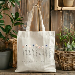 May include: A cream-colored tote bag with a floral design and text in German. The bag features a row of watercolor flowers with words like "liebe" and "freude" beneath them. The text "GALATER 5:22-23" is printed at the bottom.