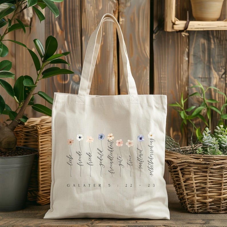 May include: A cream-colored tote bag with a floral design and text in German. The bag features a row of watercolor flowers with words like "liebe" and "freude" beneath them. The text "GALATER 5:22-23" is printed at the bottom.