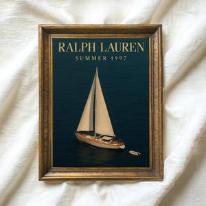 Op de afbeelding: Een ingelijste print met een zeilboot op donkerblauw water. De afbeelding is ingelijst in een goudkleurige houten lijst. De tekst "RALPH LAUREN SUMMER 1997" staat in goudkleurige letters bovenaan de afbeelding.