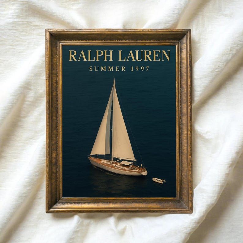 Op de afbeelding: Een ingelijste print met een zeilboot op donkerblauw water. De afbeelding is ingelijst in een goudkleurige houten lijst. De tekst "RALPH LAUREN SUMMER 1997" staat in goudkleurige letters bovenaan de afbeelding.