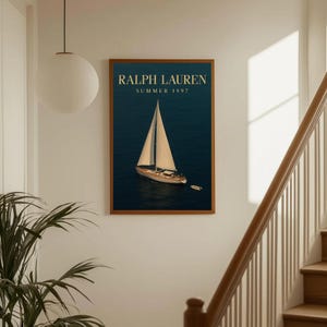 Op de afbeelding: Een ingelijste print met een zeilboot op een donkerblauwe zee. De print heeft een houten lijst en de tekst "RALPH LAUREN SUMMER 1997" in gouden letters. De afbeelding wordt weergegeven op een witte muur bij een trap.