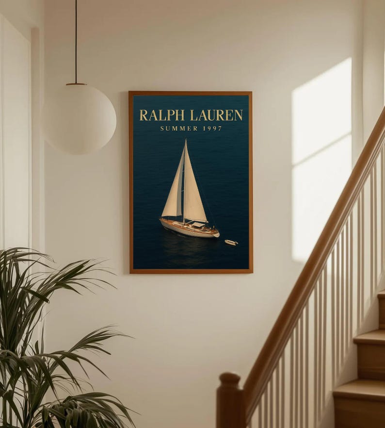 Op de afbeelding: Een ingelijste print met een zeilboot op een donkerblauwe zee. De print heeft een houten lijst en de tekst "RALPH LAUREN SUMMER 1997" in gouden letters. De afbeelding wordt weergegeven op een witte muur bij een trap.